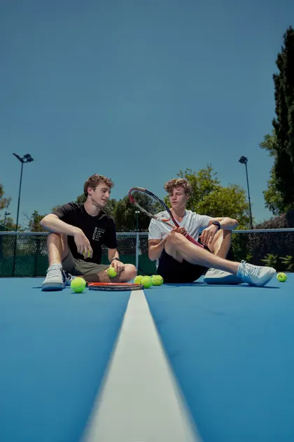 Mavi tenis kortunda raketleri ve toplarıyla oturan iki genç erkek model, güneşli gün