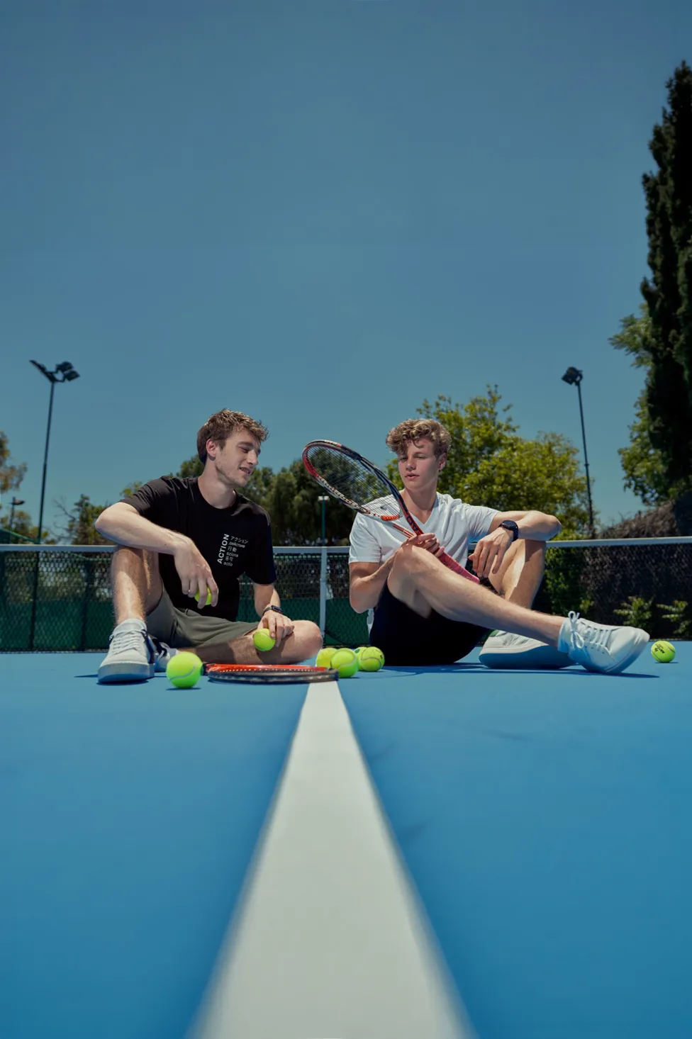 Mavi tenis kortunda raketleri ve toplarıyla oturan iki genç erkek model, güneşli gün