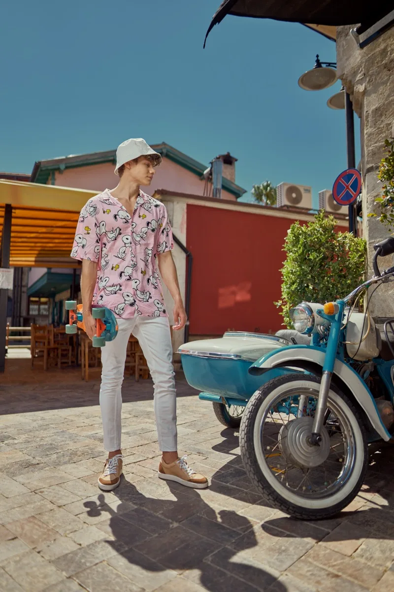 Young man in a pink patterned shirt and bucket hat holding a skateboard beside a vintage blue sidecar motorcycle
